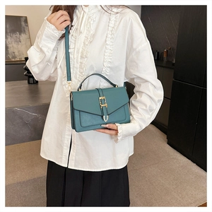 Teal leather crossbody bag with gold buckle shown on a model in a modern white outfit