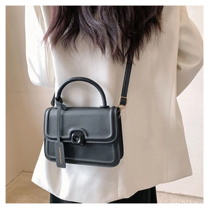 Detail of a matte black leather square bag with gold hardware and fashion tag shown on model