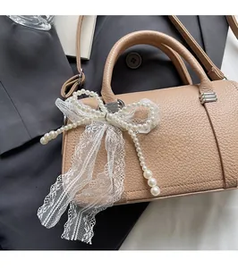 Dettaglio borsa in pelle beige con fiocco in pizzo e ciondolo di perle su sfondo grigio