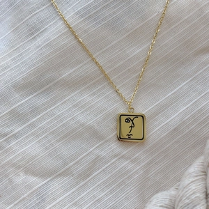 Collier carré doré minimaliste avec gravure de visage abstrait sur tissu beige texturé