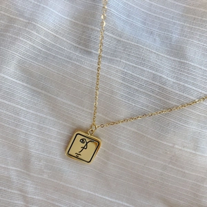 Détail du pendentif carré doré minimaliste avec contour de visage abstrait noir sur lin