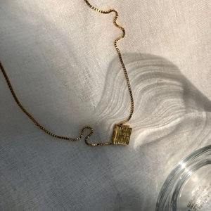 Golden necklace with cube pendant featuring geometric H-patterns on a white textured background