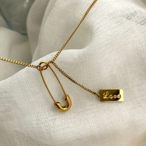 Close-up of golden safety pin and rectangular pendant with Love engraving on chain