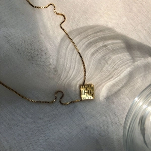 Detailed close-up of golden box chain with engraved square pendant on white fabric