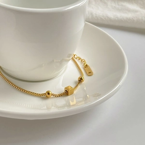 Elegant 18K gold box chain bracelet with cube and bead charms on a white ceramic cup