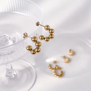 Detail of 18K gold bead C-shaped earrings hanging on glass with a soft white fabric background
