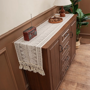 Elegant cream lace table runner with floral patterns and fringe tassels on a wooden cabinet