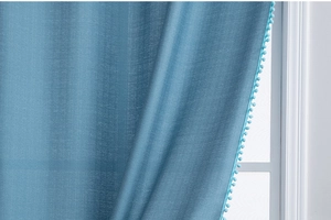 Close-up of deep sky blue curtain with decorative pom-pom trim hanging by window