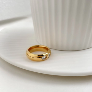 Gros plan d'une large bague dorée ornée de deux zircons ronds sur un plateau blanc