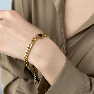 Gold Cuban chain bracelet with emerald-cut green stone worn on a female wrist