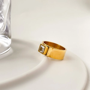 Vue détaillée d'une bague dorée large avec zircon carré sur un plateau blanc