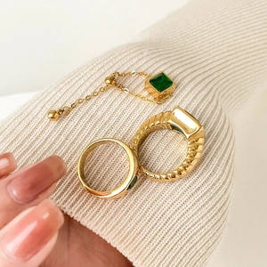 Detail of square emerald green gemstone rings and gold chain on beige knitwear texture