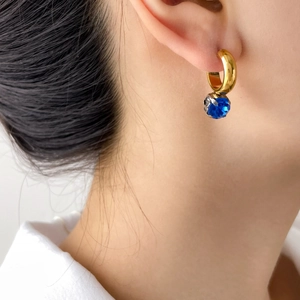 Detail of a C-shaped gold earring with a faceted blue gemstone on a model's ear