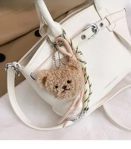 Close-up of cute light brown plush bear charm with woven strap on white leather bag