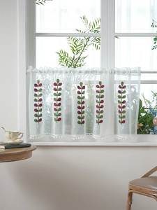 Close-up of white sheer cafe curtain with green leaf embroidery and pom-pom trim detail