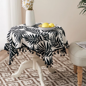 Close-up of round cotton linen tablecloth featuring black tropical leaf prints and fringe edge