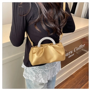 Golden pleated cloud bag with pearl handle shown on shoulder in a cafe setting