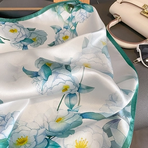 Elegant mulberry silk scarf with camellia print and green hem, next to a leather handbag