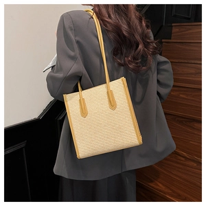 Straw tote bag with yellow leather trim shown on shoulder in a minimalist indoor setting