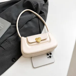Dettaglio borsa a tracolla color crema con fibbia a cuore dorata accanto a uno smartphone