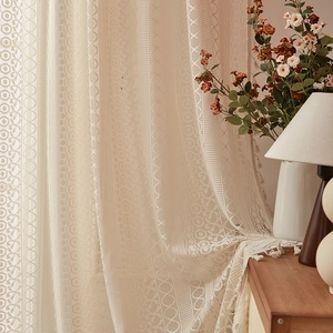 Detail of cream lace curtain with intricate patterns and fringe tassels in cozy room