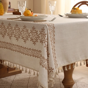 Beige linen tablecloth with brown floral embroidery and fringe tassels on a wooden table
