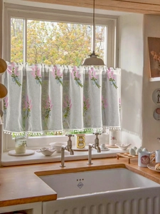 Detailed embroidery of pink flowers and blue ribbons on a white pom-pom cafe curtain