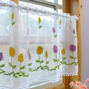 Sheer white cafe curtain with tulip embroidery and pom-pom trim on a wooden window