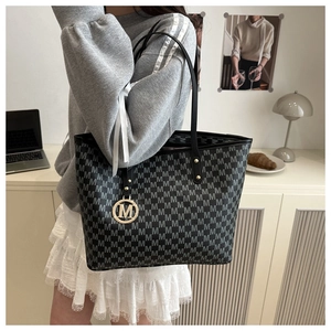 Close-up of black tote bag with gray monogram pattern and golden M-shaped metal pendant