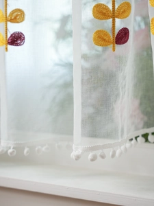 Detailed macro view of white sheer mesh curtain featuring yellow floral embroidery and pom-poms