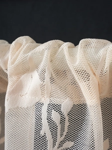 Dettaglio macro della tenda in pizzo beige con trama a rete fine e motivi floreali