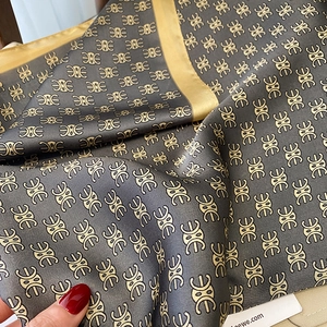 Detailed view of the silk scarf featuring golden Anagram patterns on a dark grey fabric