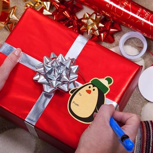 Close-up of a hand writing on a penguin Christmas gift tag with festive wrapping paper