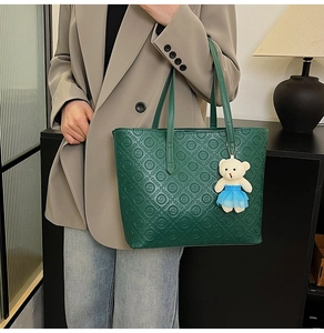 Detail of dark green tote bag with embossed logo and white plush bear pendant accessory