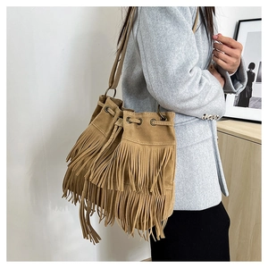 Brown suede bucket bag with long tassels and drawstring worn by a model in a home setting