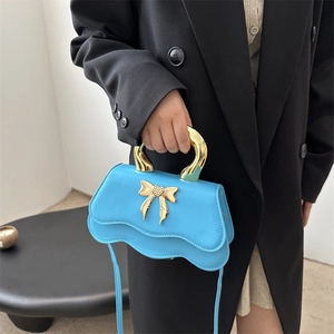 Blue crossbody bag with gold bow handle shown on model in a minimalist black suit