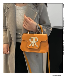 Detail of a bright orange leather crossbody bag featuring a bold golden logo on gray coat