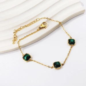 Détail de bracelet doré avec pierres carrées vert émeraude sur support blanc ondulé