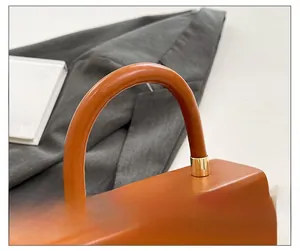 Detail of a brown glossy leather handbag featuring a curved handle with gold metal accents
