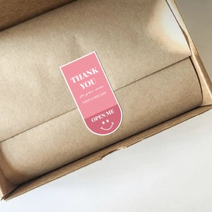 Pink rectangular thank you stickers with smiley faces on a kraft shipping box