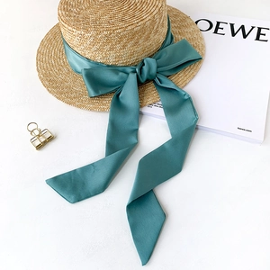 Flat top straw sun hat with elegant wide dark cyan satin ribbon bow on white surface