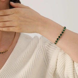 Goldenes Armband mit smaragdgrünen Kristallen an einer Frau in weißem Strickpullover