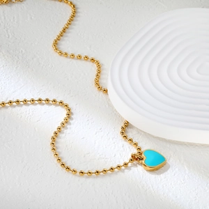 Détail du collier à billes dorées avec pendentif cœur émaillé turquoise sur socle blanc