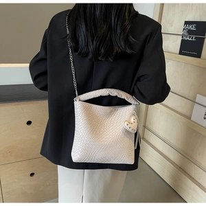 Detail der cremefarbenen gewebten Beuteltasche mit Kettengurt und Herzanhänger am Modell