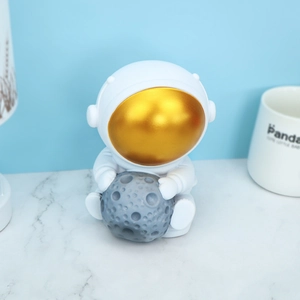 Creative astronaut piggy bank in white and gold finish holding a grey moon on marble surface