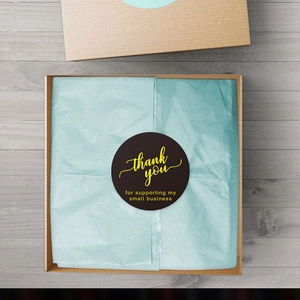 Black round 'thank you' stickers with gold foil text on blue tissue paper in a kraft box