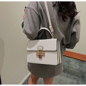 Detail einer weißen Lederhandtasche mit goldenem Bienenverschluss und Perlenverzierung