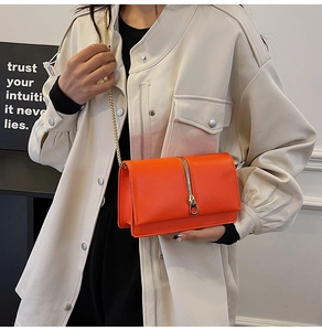 Close-up of bright orange crossbody bag with gold chain strap and zipper detail on model