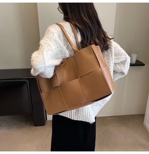 Detail of a large brown woven tote bag worn by a model in a minimalist indoor setting