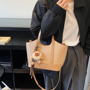 Khaki Beuteltasche mit Lederstruktur und Löwen-Anhänger vor einem Bücherregal